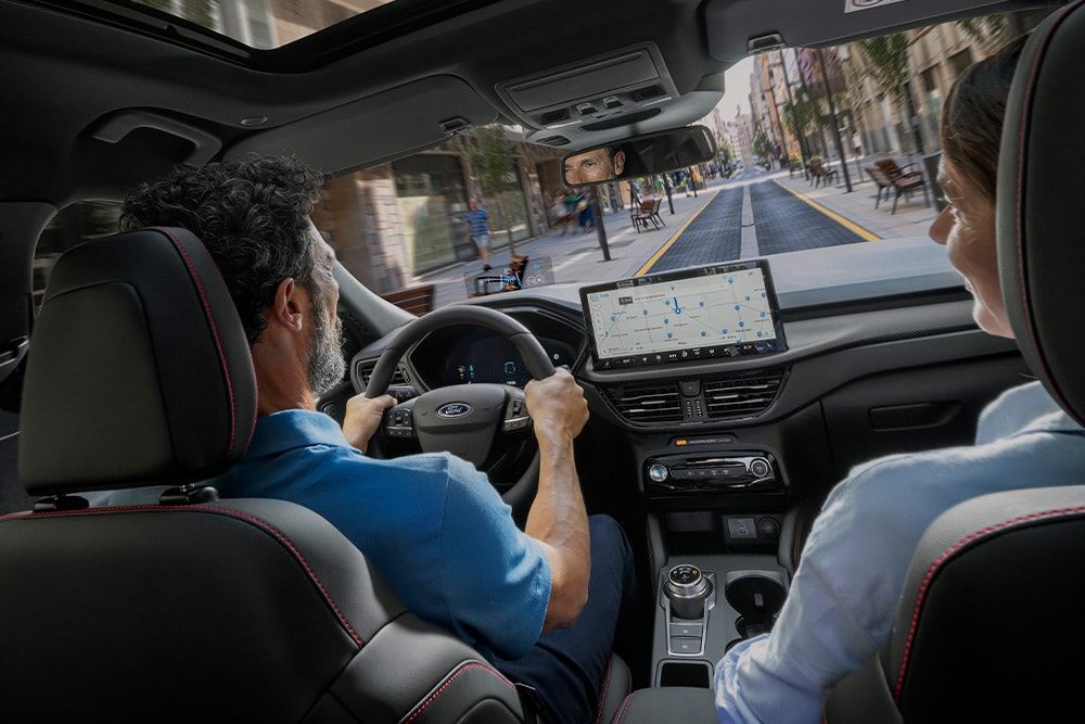 Zwei Personen sitzen in einem Ford Kuga, fahren in der Stadt auf einer Straße und folgen der Navigation auf dem Touchscreen.
