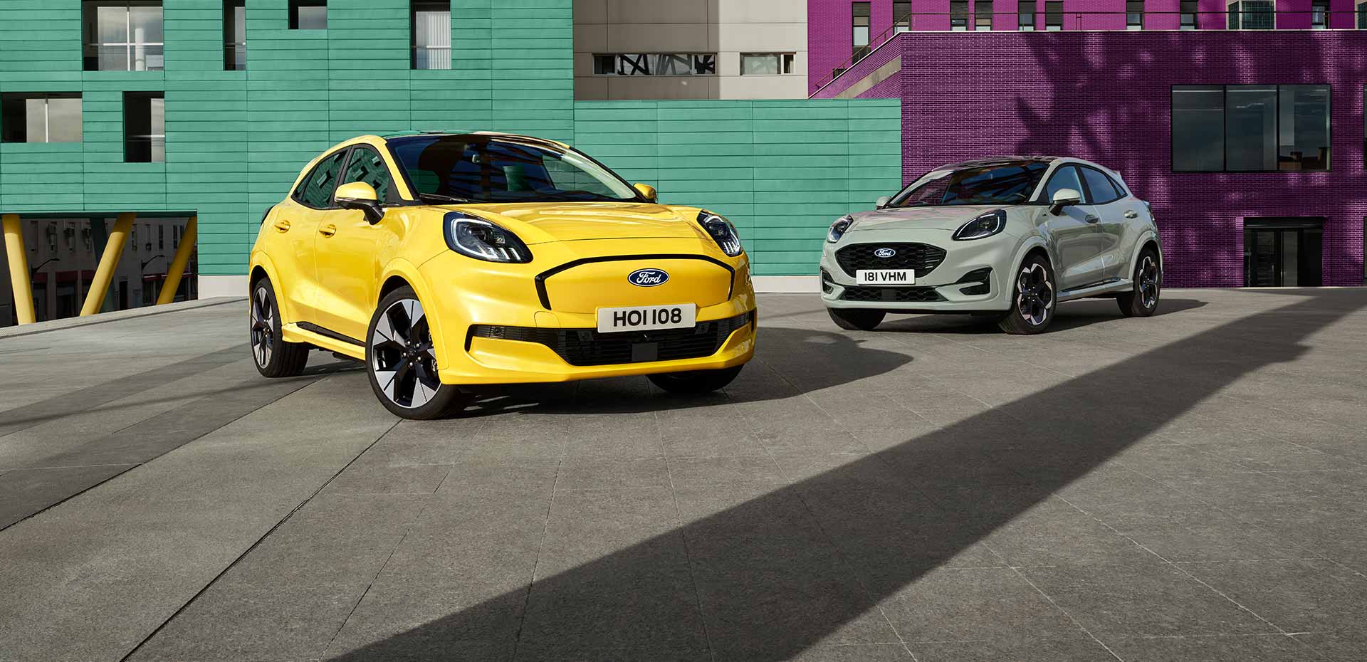 Ford Puma Gen-E in Gelb und Ford Puma in Grau. Dreiviertelansicht, stehend vor einer bunten Wand.