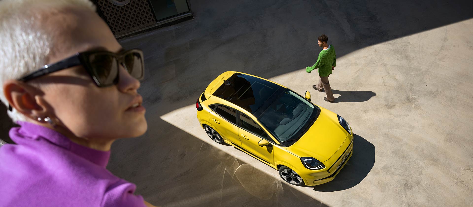 Ford Puma Gen-E in Gelb. Ansicht von oben, parkend auf einer Fläche. Person nähert sich dem Fahrzeug.
