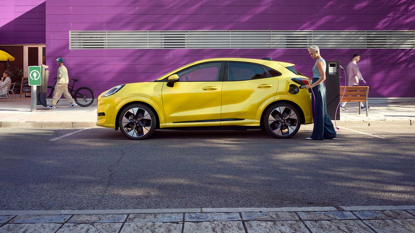 Ford Puma Gen-E in Gelb. Seitenansicht, parkend an einer Ladestation. Person verbindet das Ladekabel mit dem Fahrzeug.