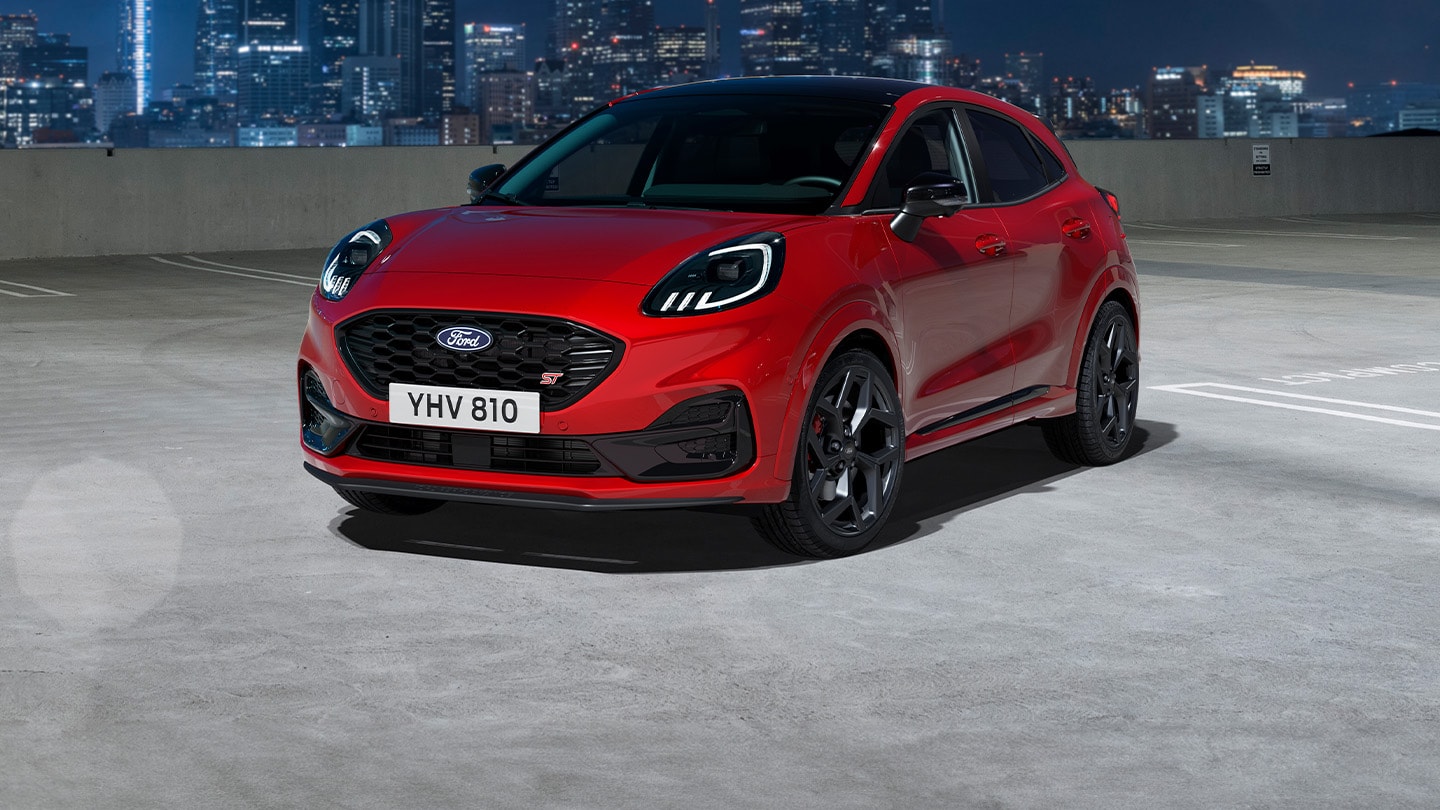 Ford Puma in Rot. Dreiviertelansicht, stehend auf einem Parkplatz bei Nacht. Beleuchtete Gebäude im Hintergrund.