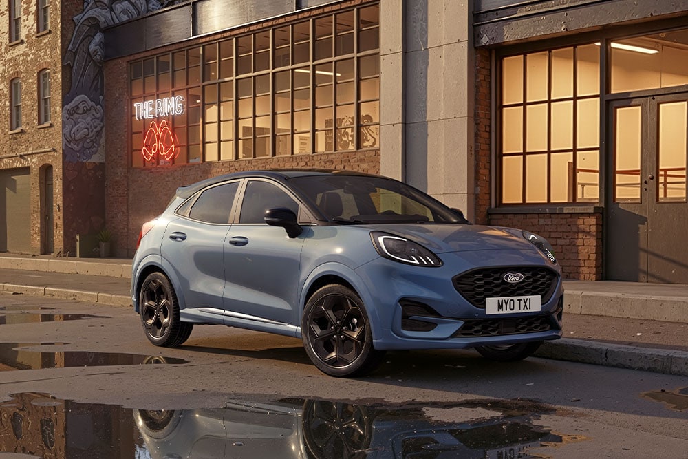 Ein blauer Ford Puma parkt bei Sonnenuntergang auf einer städtischen Straße mit Pfützen, die die Gebäude widerspiegeln.