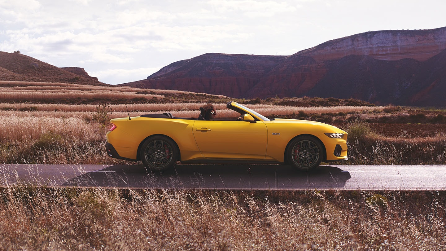 Ford Mustang in Gelb. Seitenansicht, fahrend auf einer Straße. Frau sitzt im Fahrersitz.