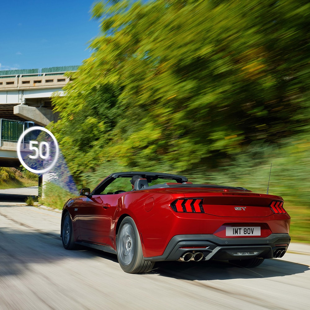 Ford Mustang in Rot. Dreiviertelansicht, fahrend auf einer Straße mit Bäumen im Hintergrund.