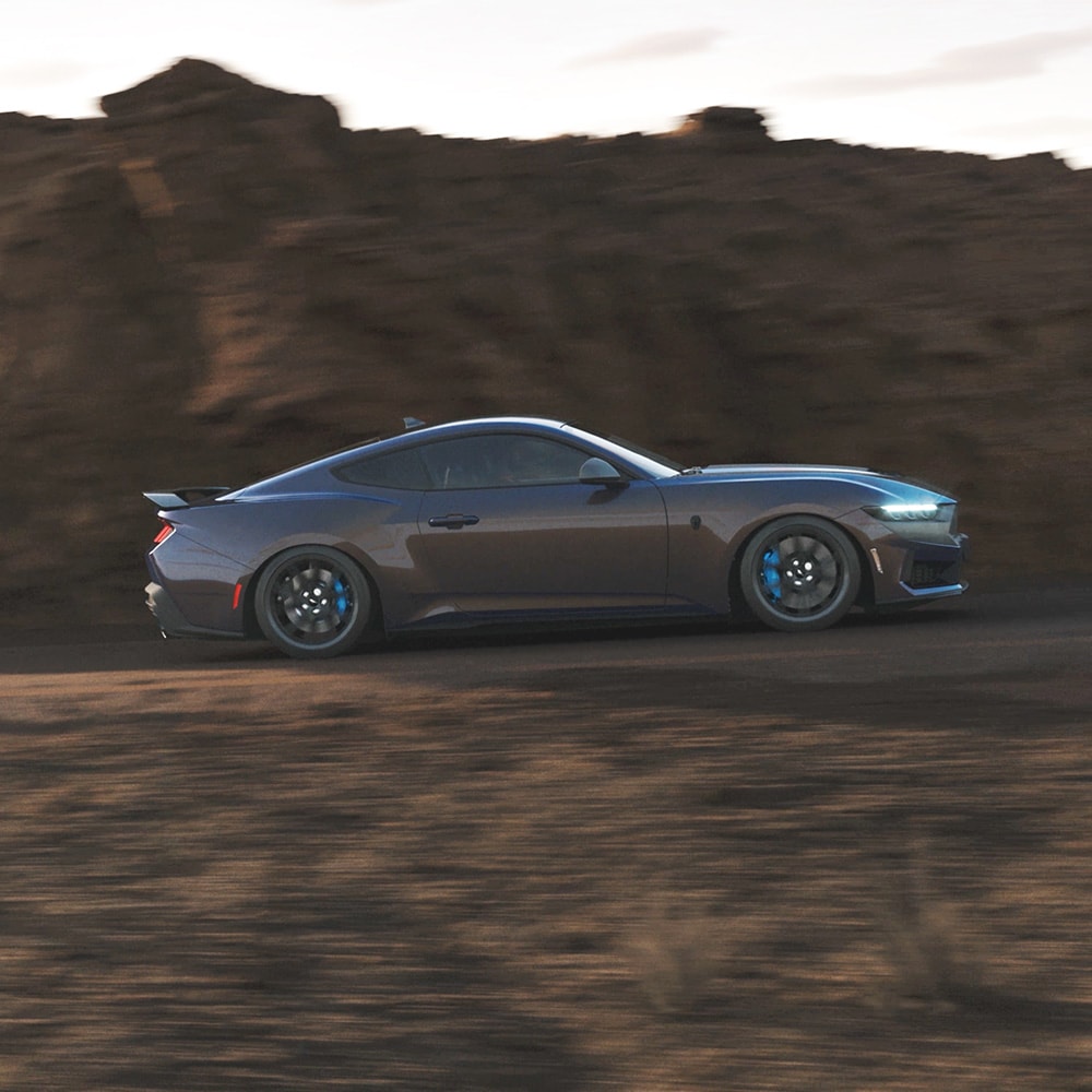 Ford Mustang in Blau. Seitenansicht, fahrend auf einer Straße, Felsen im Hintergrund.