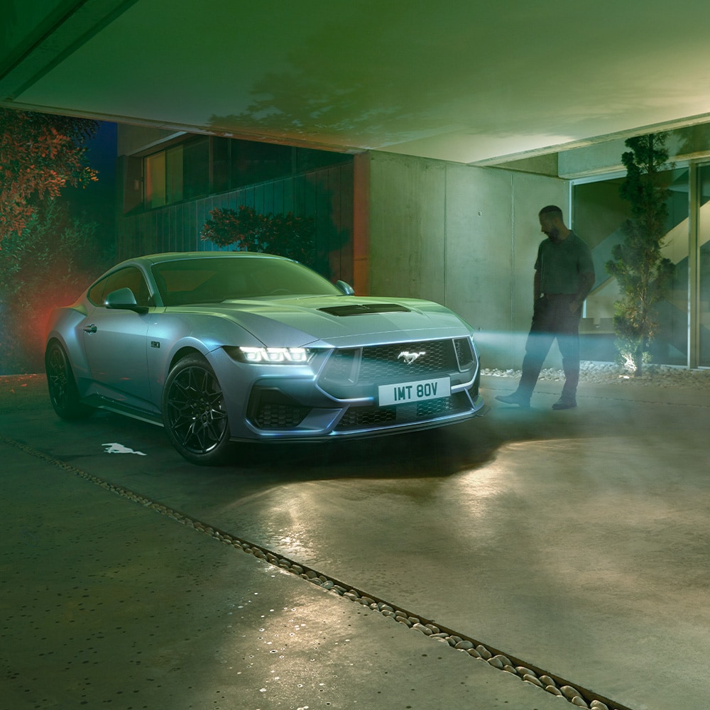 Ford Mustang in Blau. Frontansicht, parkt mit eingeschalteten Scheinwerfern vor einem Wohnhaus bei Nacht. Mann danebenstehend.