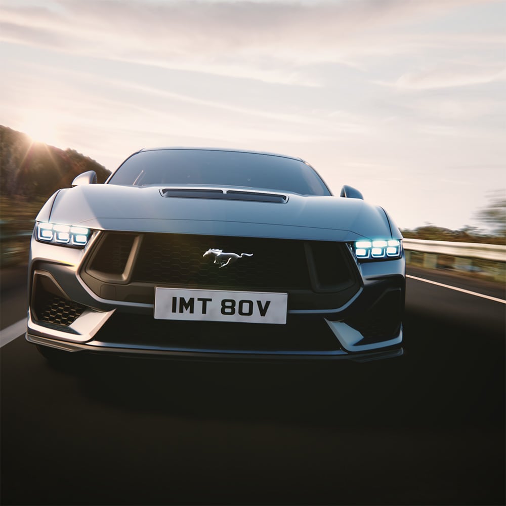Ford Mustang in Blau. Frontansicht, fahrend auf einer Straße mit Bergen im Hintergrund.