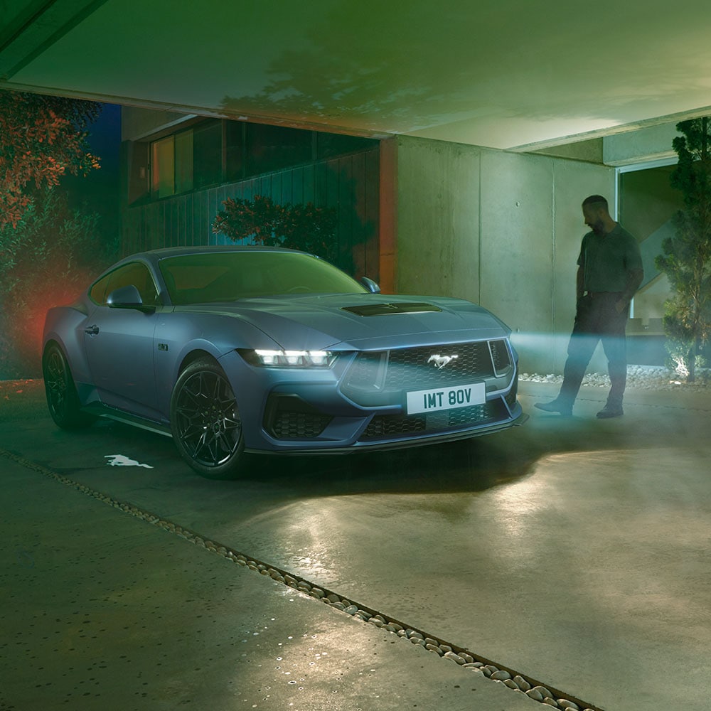 Ford Mustang in Blau. Frontansicht, parkt mit eingeschalteten Scheinwerfern vor einem Wohnhaus bei Nacht. Mann danebenstehend.