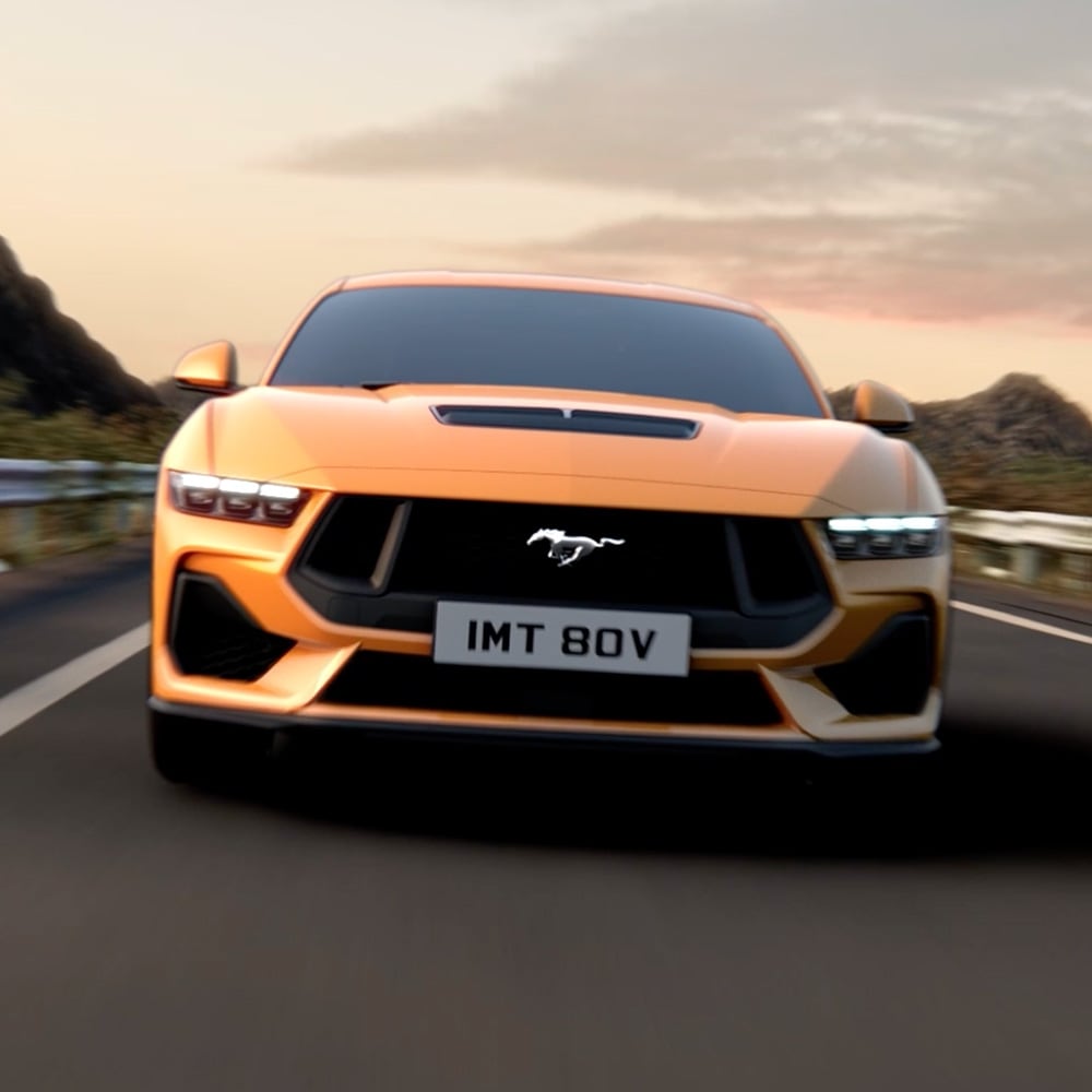 Ford Mustang in Blau. Frontansicht, fahrend auf einer Straße mit Bergen im Hintergrund.