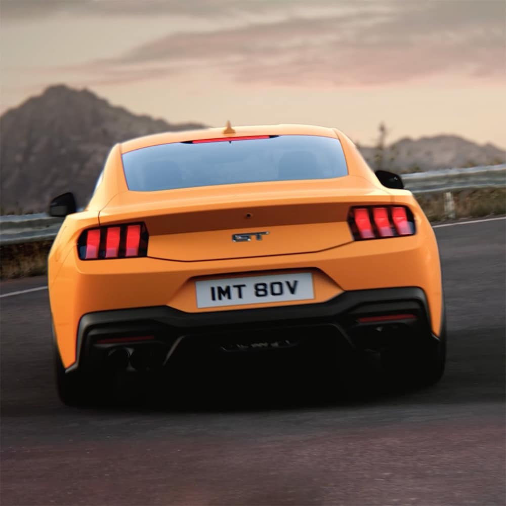 Ford Mustang in Blau. Dreiviertelansicht, fahrend auf einer unbefestigten Straße bei Sonnenuntergang.