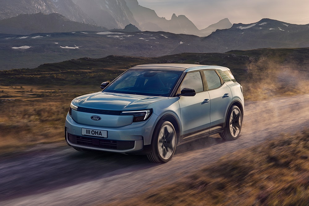 Ford Explorer in Blau. Dreiviertelansicht, fahrend auf einer Straße bei Sonnenuntergang. Berge im Hintergrund.