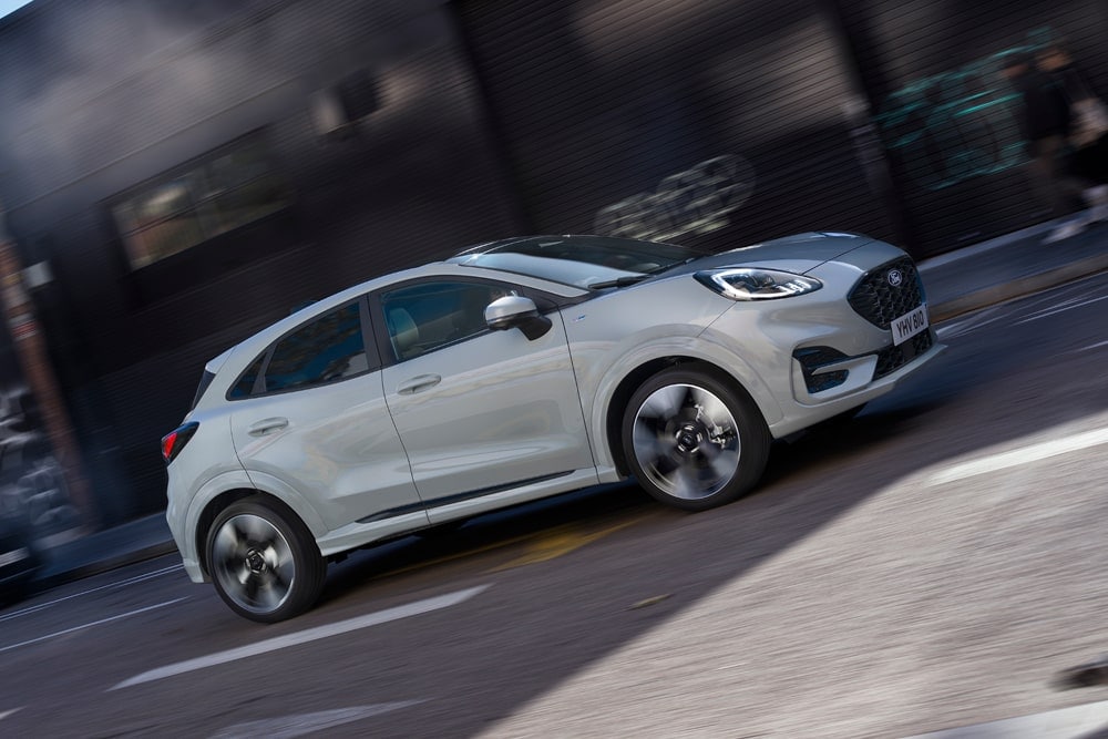 Ford Puma Gen-E in Silber aus dem Sortiment der Ford Kleinwagen. Fährt in einer Stadt eine Straße entlang.