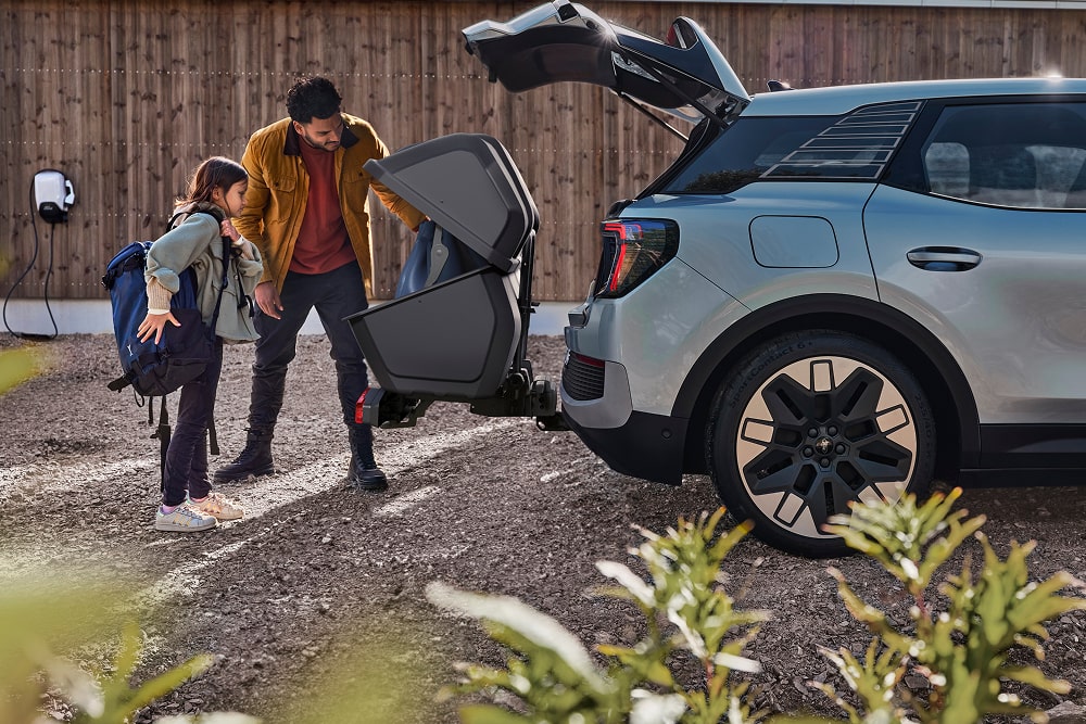 Eine Person und ein Kind stehen am geöffneten Kofferraum eines blauen Ford Explorer SUV und laden einen zusammengeklappten Kinderwagen in eine Gepäckbox ein. 