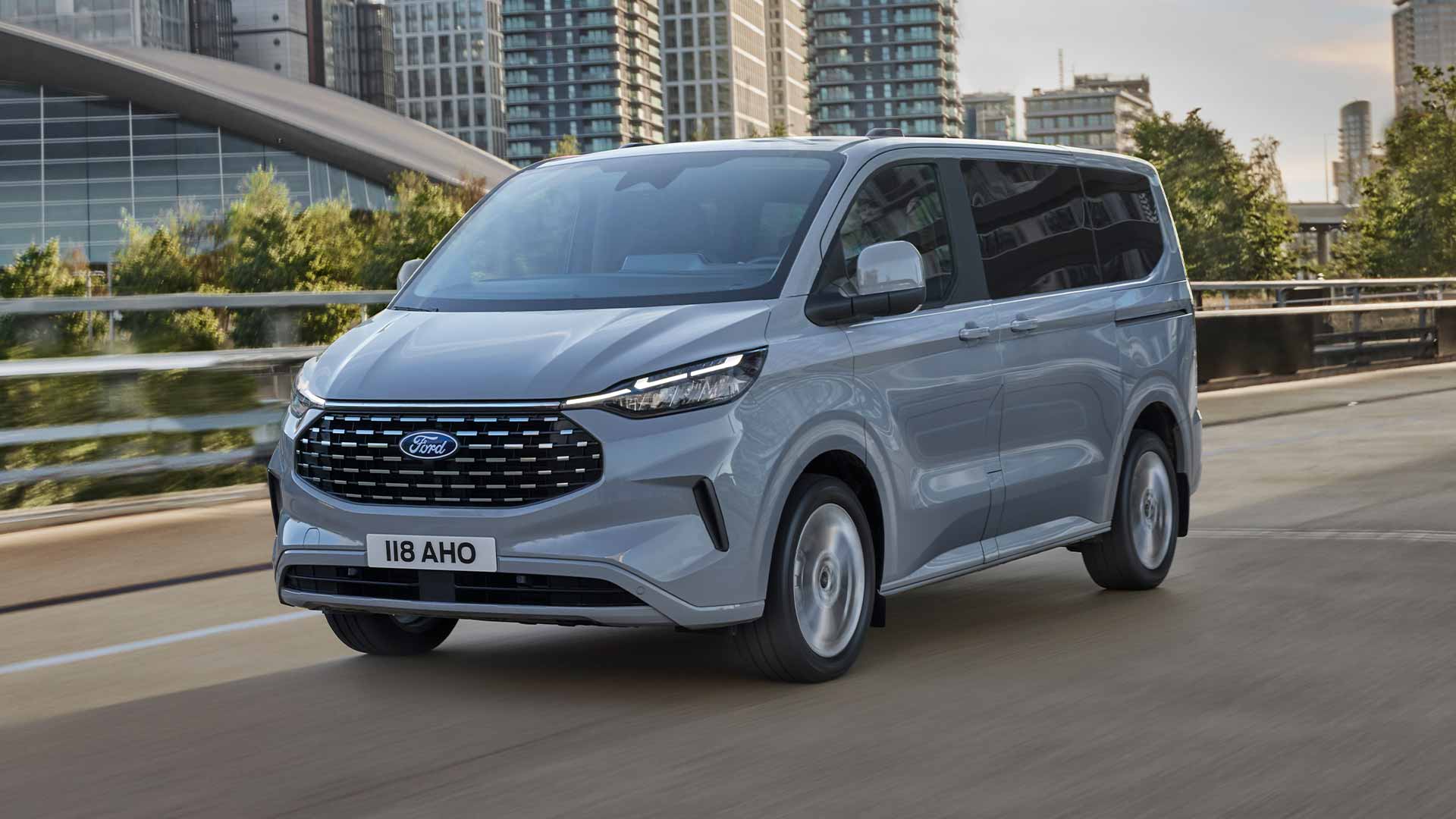 Ford Tourneo Custom in Silber. Dreiviertelansicht, fahend auf einer Straße, Hochhäuser im Hintergrund.