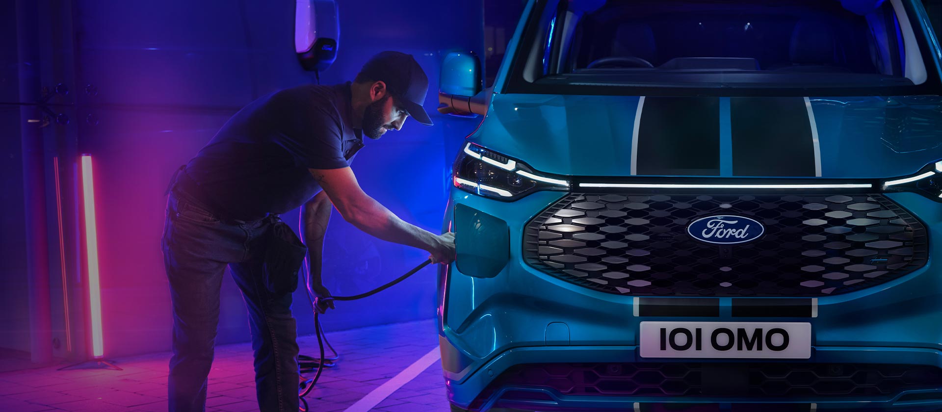 Ford E-Tourneo Custom in Blau. Frontansicht, Mann schließt Ladekabel an das Fahrzeug an.
