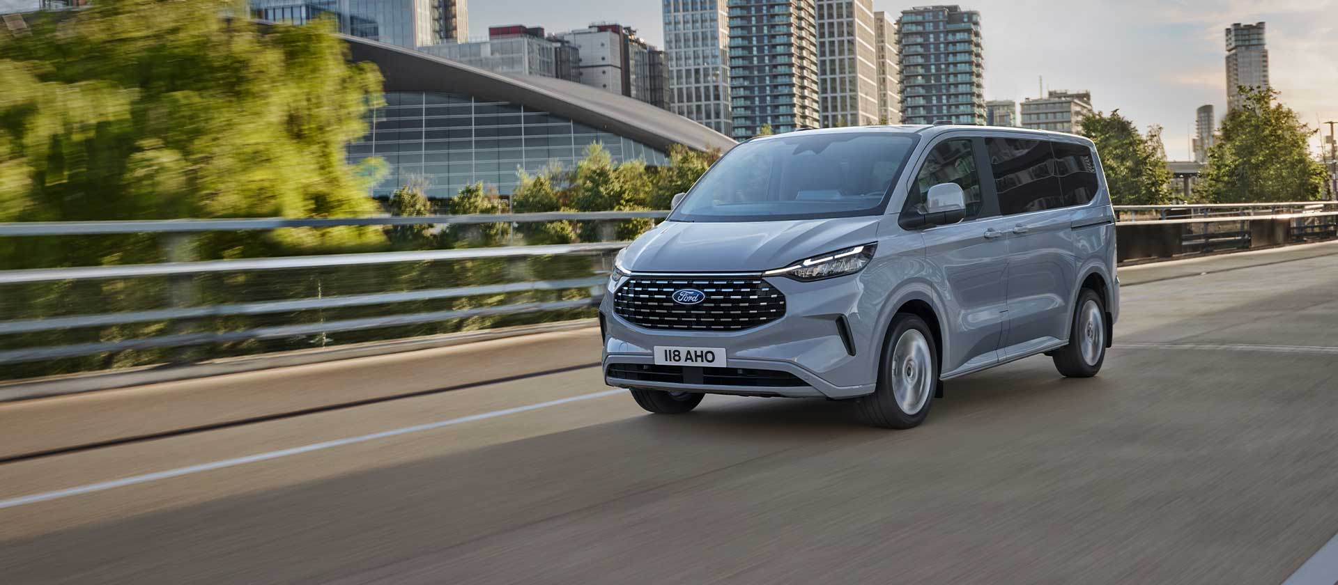 Ford Tourneo Custom in Silber. Dreiviertelansicht, fahrend auf einer Straße, Hochhäuser im Hintergrund.