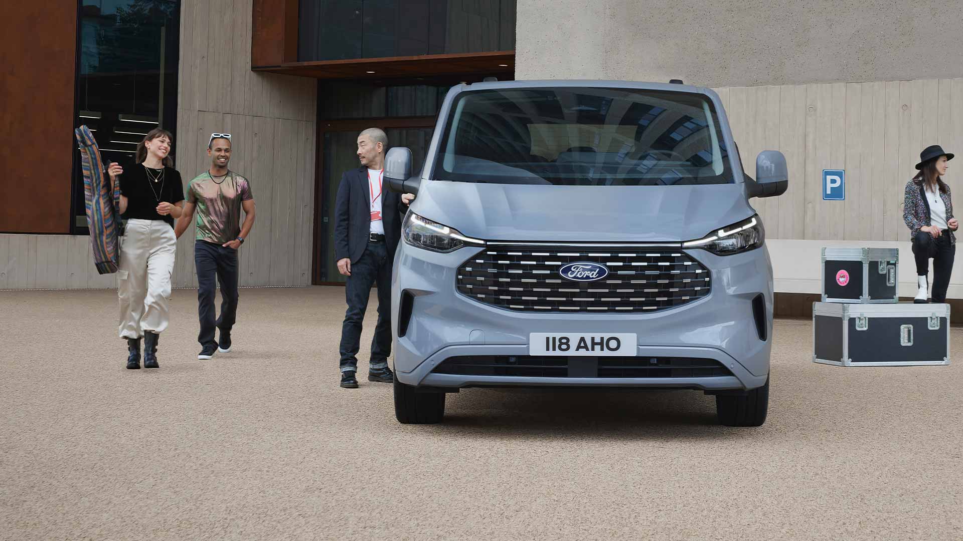 Ford Tourneo Custom in Grau. Frontansicht, auf einem Parkplatz. Mehrere Personen stehen neben dem Fahrzeug.