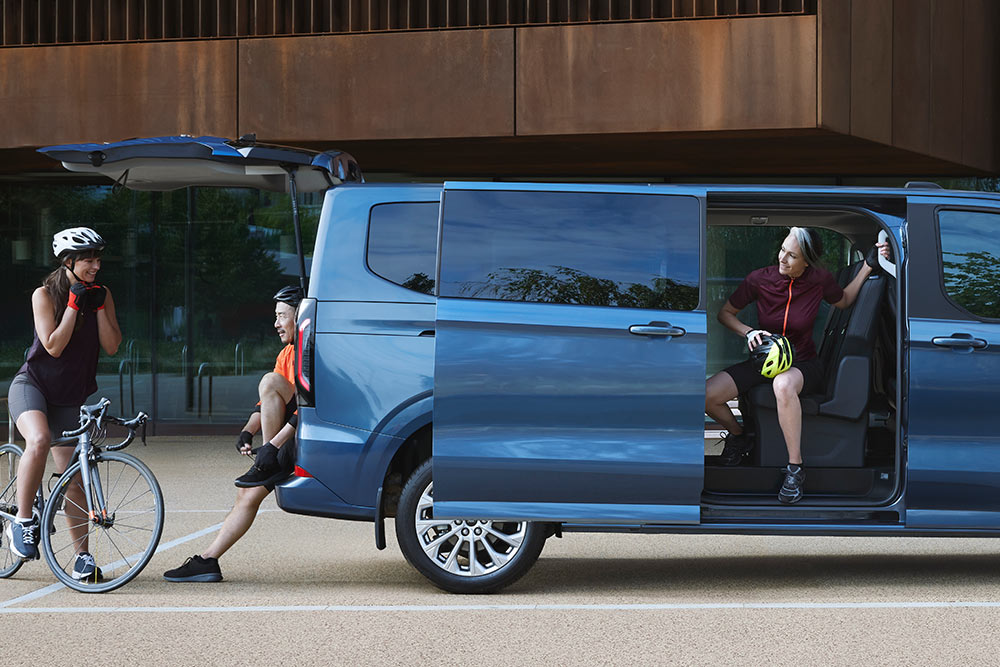 Ford Tourneo Custom in Blau. Seitenansicht, stehend vor einem Haus mit geöffneter Seitentür.
