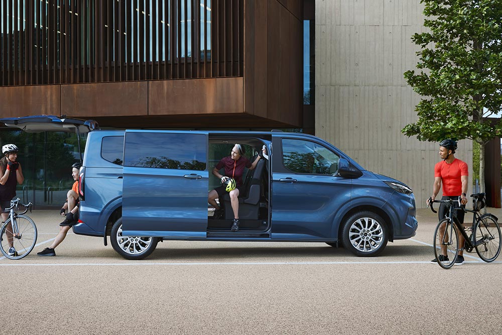 Ford Tourneo Custom in Blau. Seitenansicht, stehend vor einem Haus mit geöffneter Seitentür.