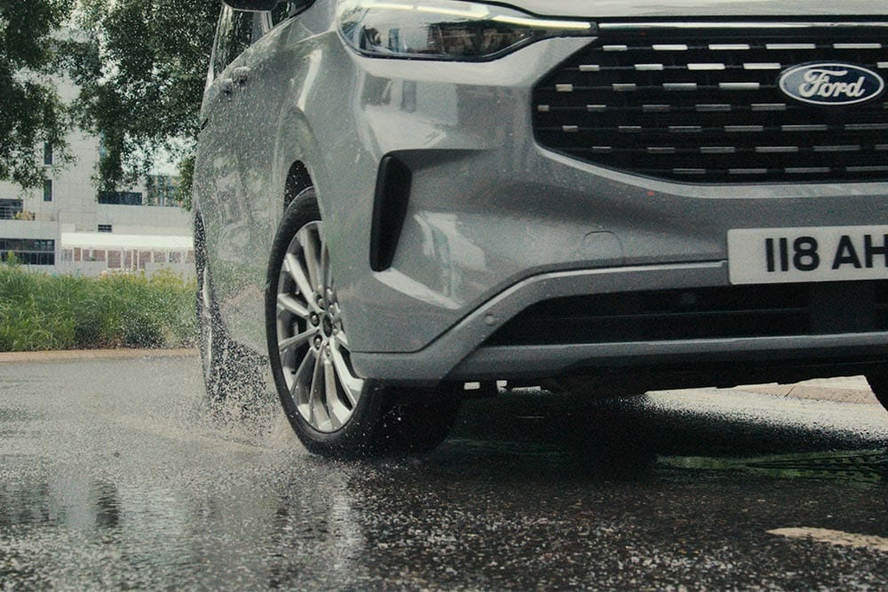 Ford Tourneo Custom in Grau. Detailansicht des rechten Vorderrads, Scheinwerfer und Kühlergrill.