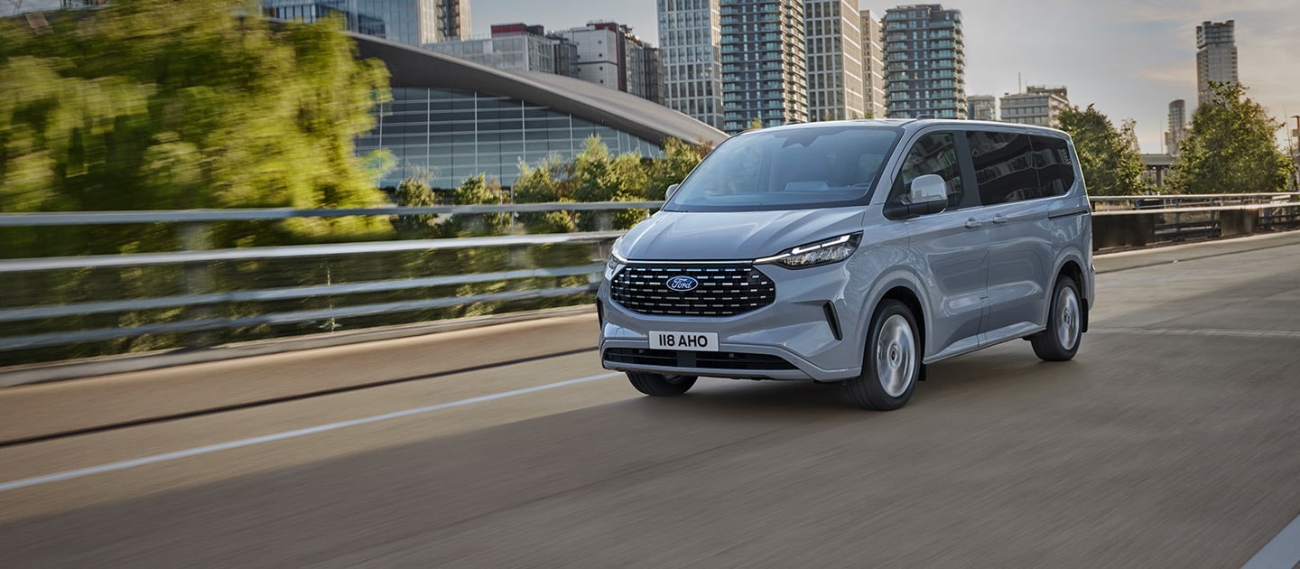 Ford Tourneo Custom in Silber. Dreiviertelansicht, fahrend auf einer Straße, Hochhäuser im Hintergrund.