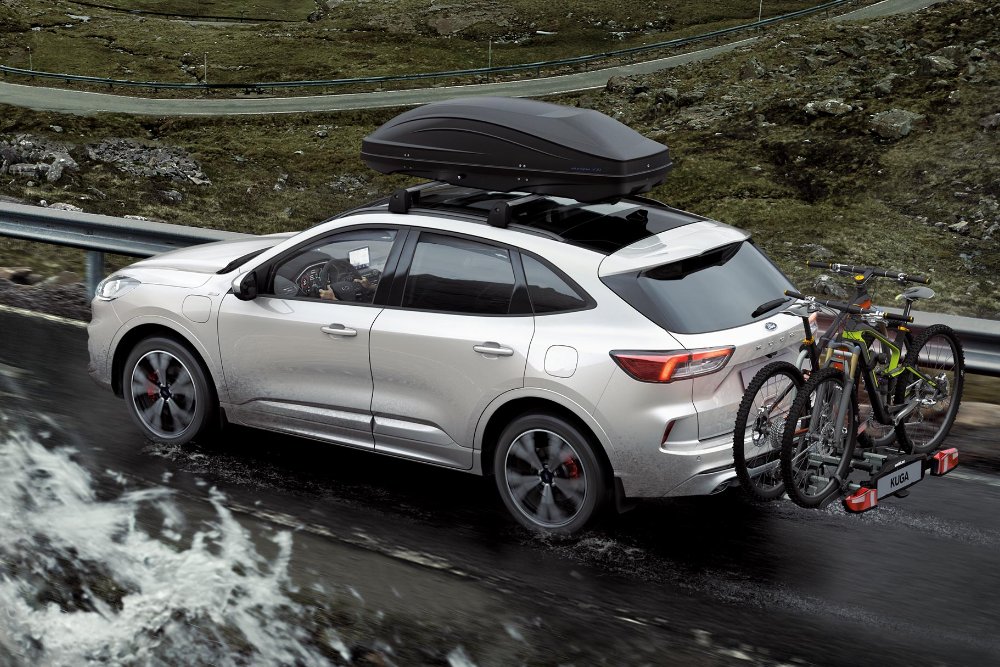 Ford Kuga in silber fahrend auf Straße in den Bergen mit Hecktrager und Fahrradern. Dreiviertelansicht hinten.