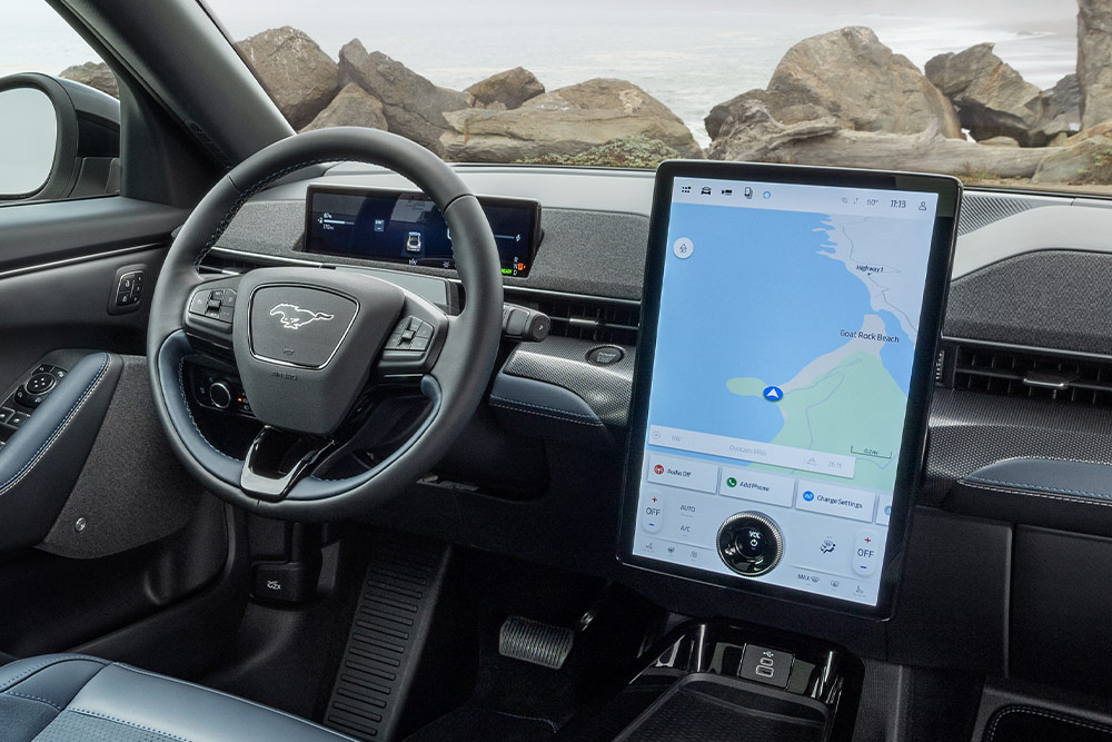 Innenansicht eines Ford Mustang Mach-E mit Armaturenbrett, Lenkrad und großem Touchscreen mit Karte, geparkt an einer felsigen Küste.
