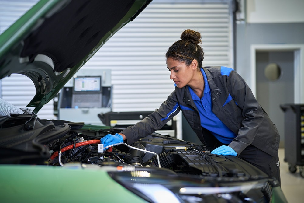 Technikerin mit blauen Handschuhen arbeitet am Motor eines grünen Autos bei geöffneter Motorhaube.