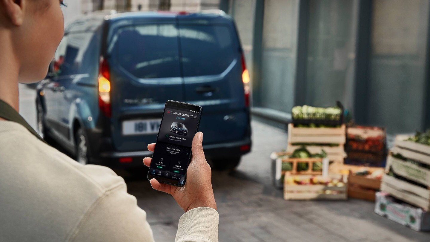 FordPass App – Frau hält ein Mobiltelefon und Ford Transit Connect in Blau. Heckansicht, parkend in einer Lagerhalle