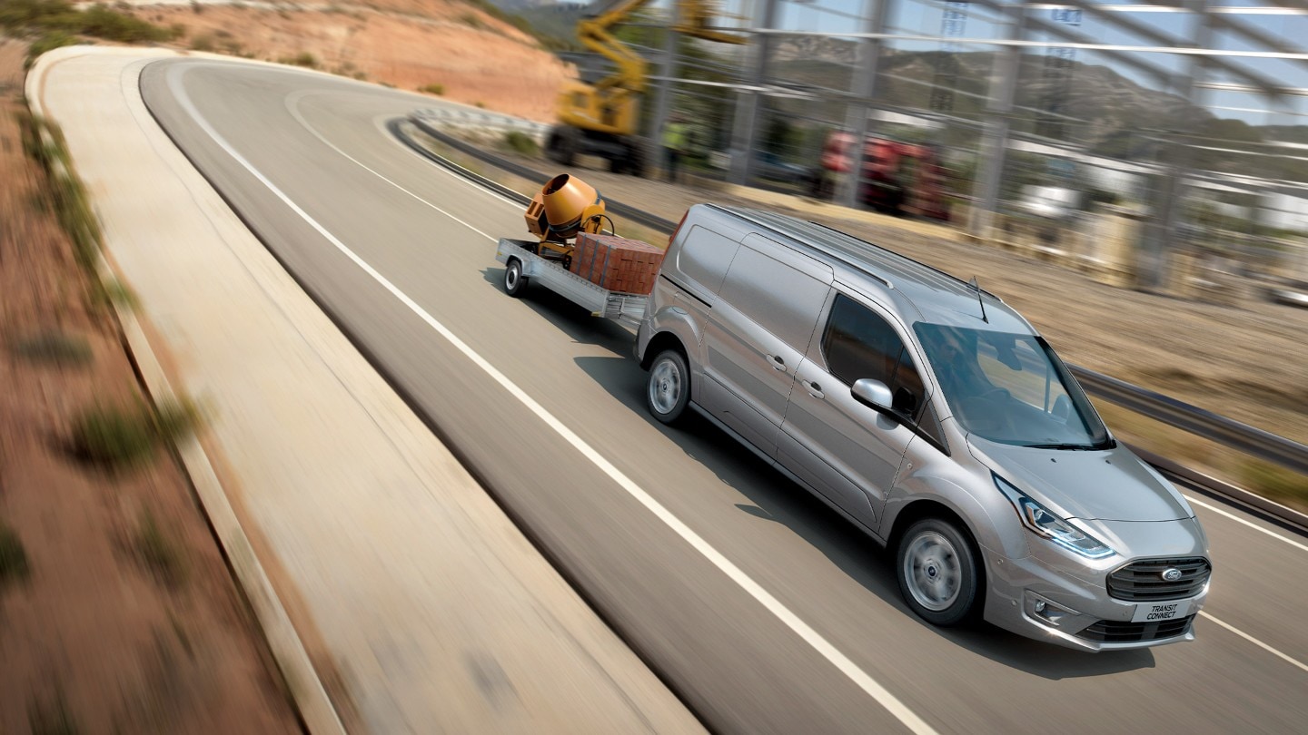 Ford Transit Connect in Silber. Dreiviertelansicht, transportiert einen Anhänger bergab auf einer Landstraße