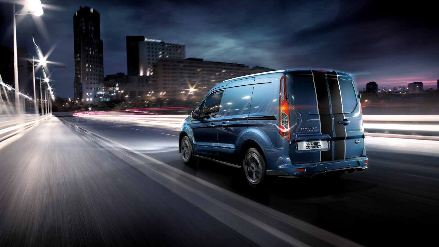 Ford Transit Connect in Blau. Dreiviertelansicht von hinten, fahrend durch eine Großstadt bei Nacht 