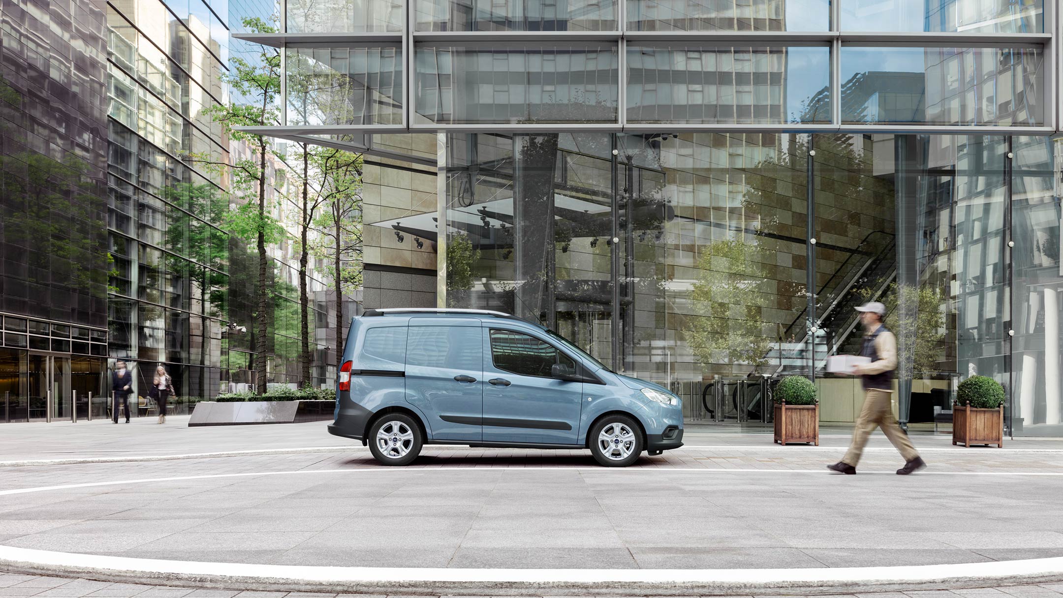 Ford Transit Courier in Blau. Seitenansicht, parkt vor einem modernen Gebäude mit Glasfassade
