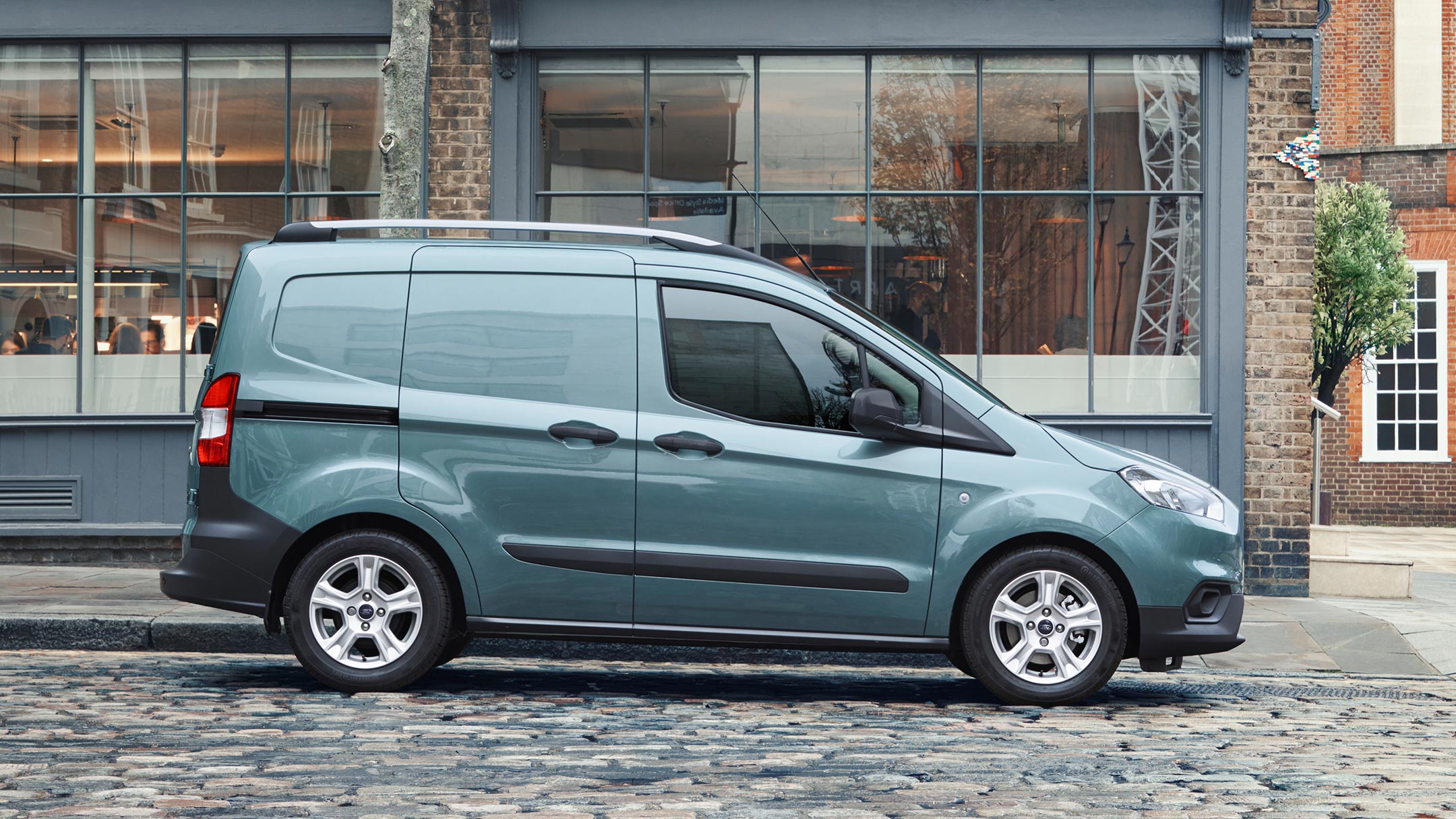 Ford Transit Courier in Blau. Seitenansicht, parkend vor einem modernen Gebäude