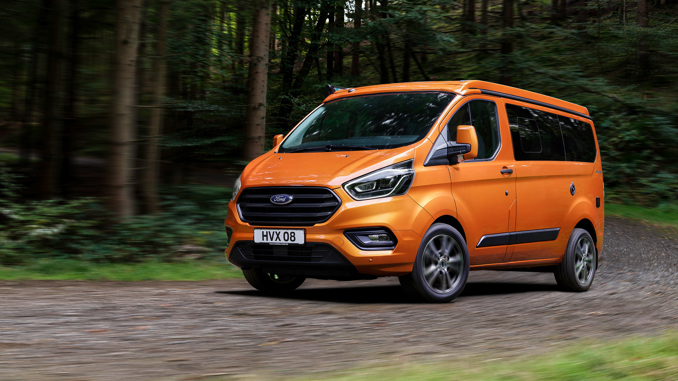 Ford Transit Custom Nugget in Orange mit Aufstelldach. Dreiviertelansicht, fahrend im Wald