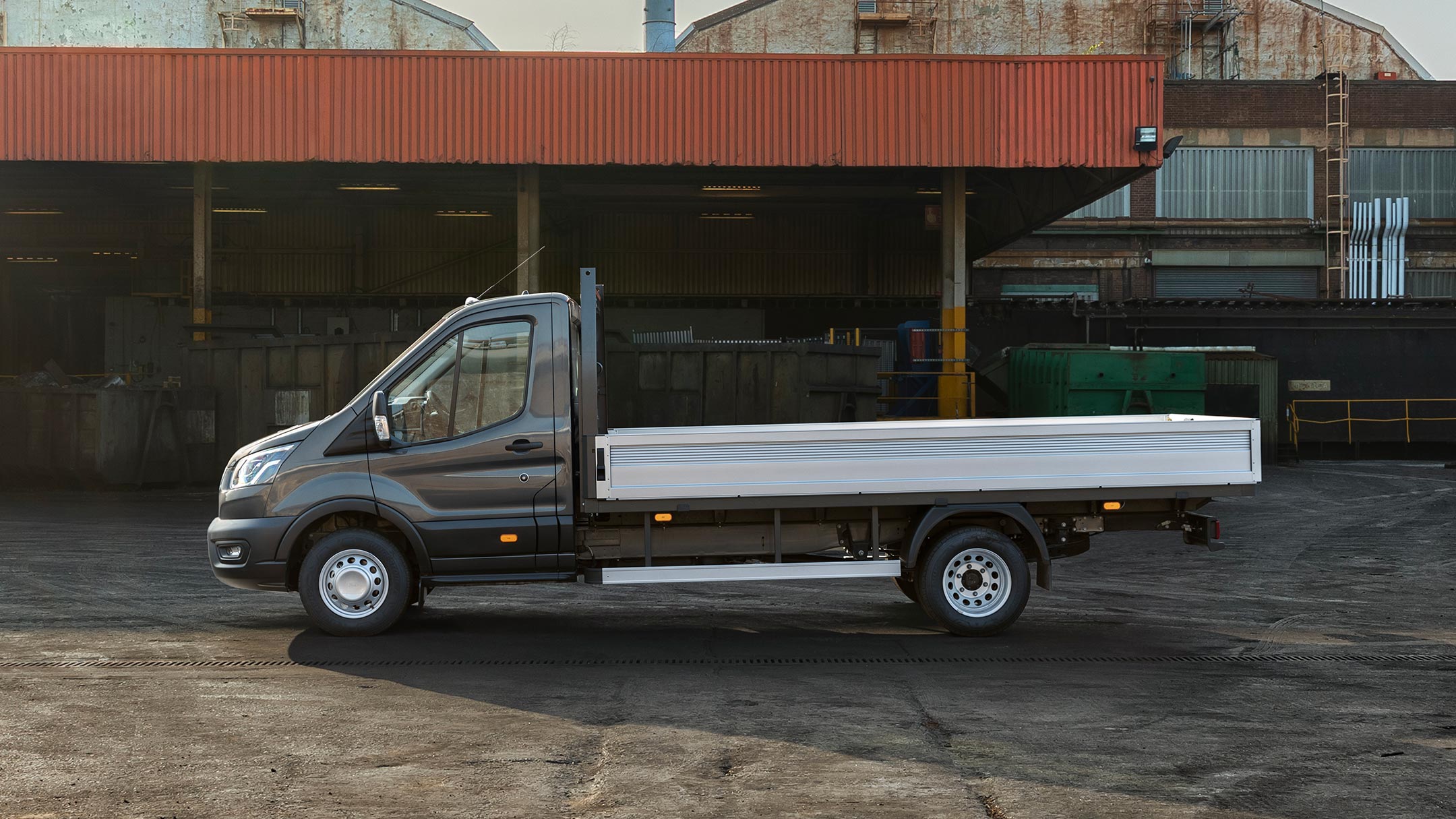 Ford Transit 5 Tonnen Fahrgestell in Schwarz. Seitenansicht, parkend auf einem Betriebsgelände neben einer Überdachung mit Paletten