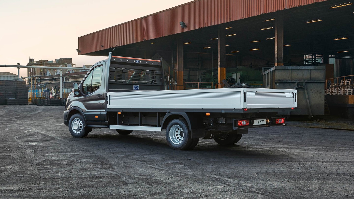 Ford Transit 5 Tonnen Pritschenwagen in Schwarz. Dreiviertelansicht von hinten, parkend auf einem Betriebsgelände neben einer Überdachung mit Paletten