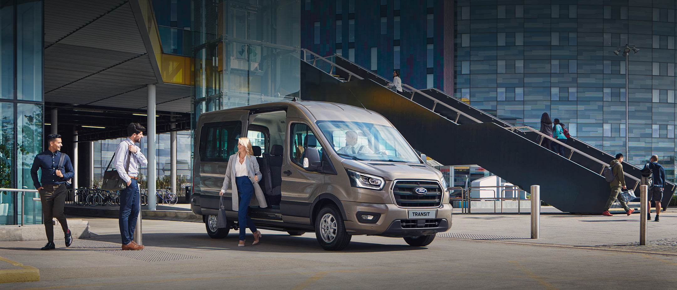 Ford Transit Bus in Silber. Dreiviertelansicht, vor einem Glasgebäude geparkt