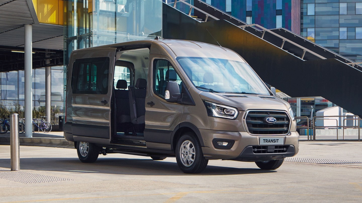 Ford Transit Bus in Silber. Dreiviertelansicht, parkend vor modernem Gebäude mit geöffneter Schiebetür 