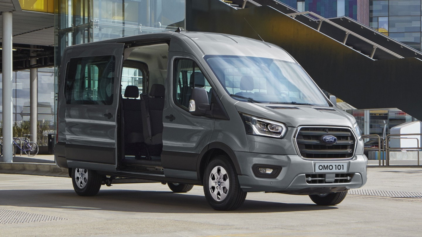 Ford Transit Bus in Silber. Dreiviertelansicht, parkend vor modernem Gebäude mit geöffneter Schiebetür.