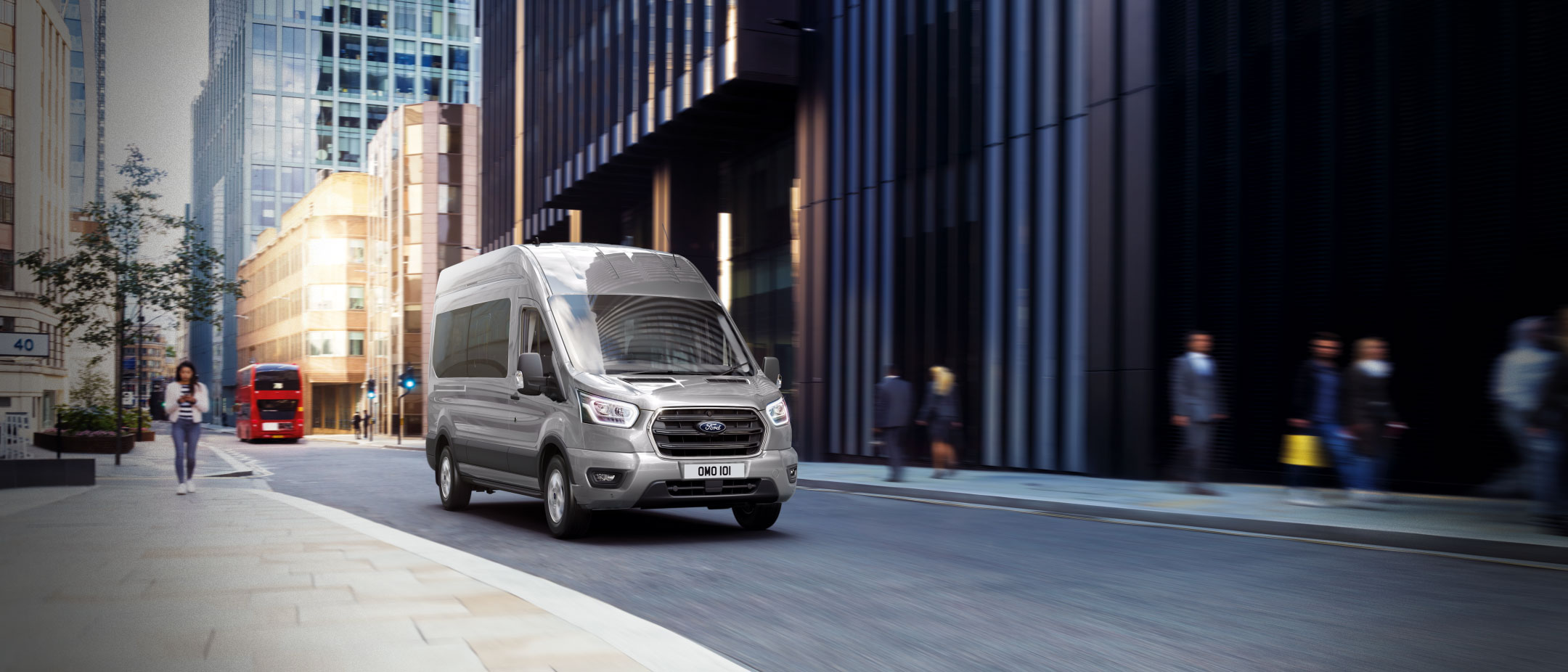 Ford Transit Bus in Silber. Dreiviertelansicht, fahrend durch die Innenstadt.