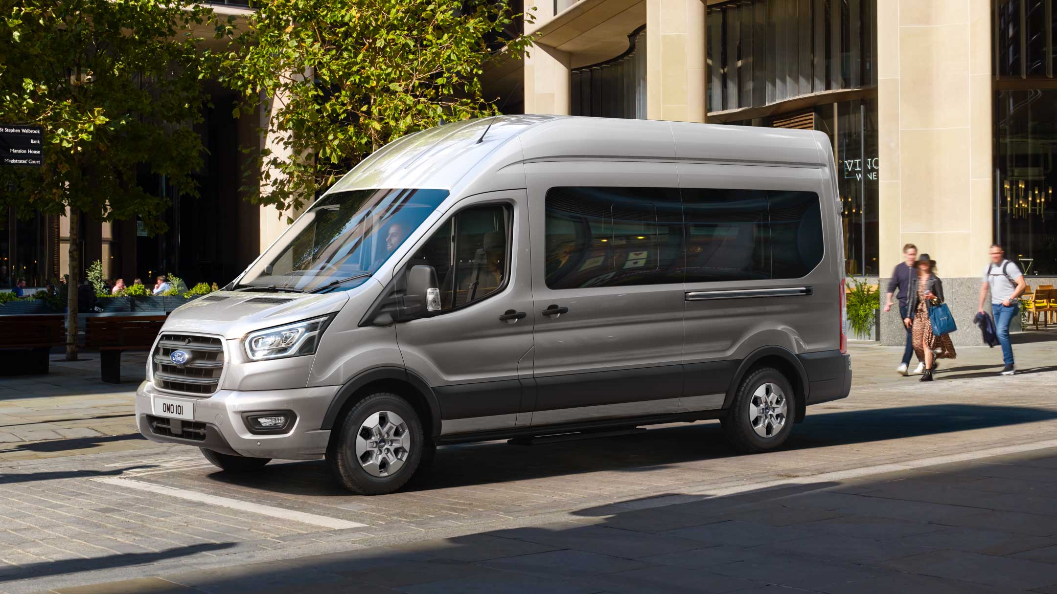 Ford Transit Bus in Silber. Seitenansicht, parkend vor einem Restaurant.