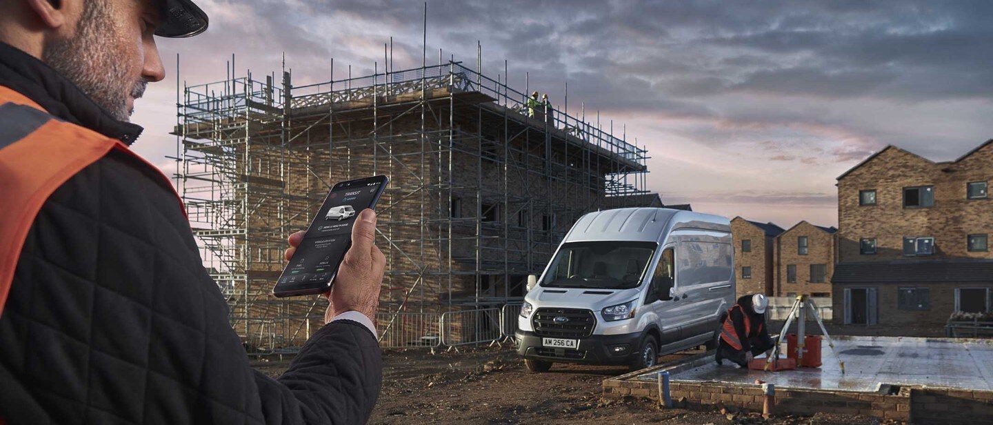 Ford Transit in Silber. Frontansicht, auf einer Baustelle. Mann sieht auf sein Smartphone mit geöffneter FordPass App