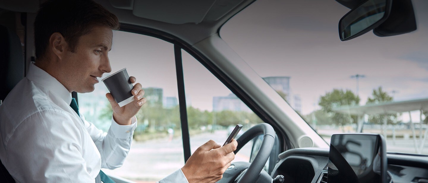 Mann sitzt in einem Ford und trinkt Kaffee während er auf sein Smartphone schaut