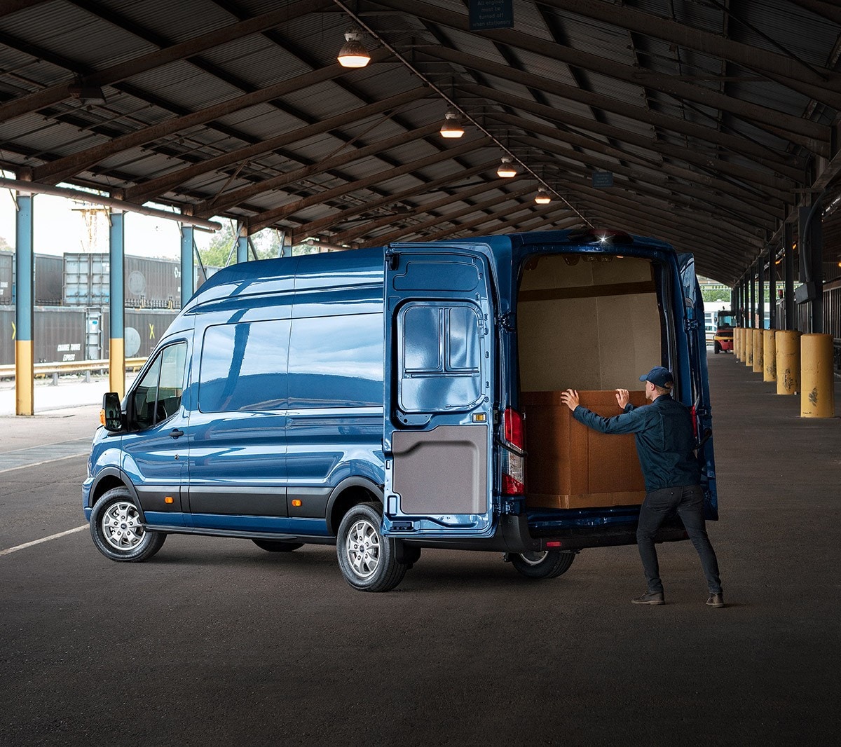 Ford Transit Kastenwagen in Blau. Dreiviertelansicht von hinten, parkend unter einem Vordach mit weit geöffneten Hecktüren