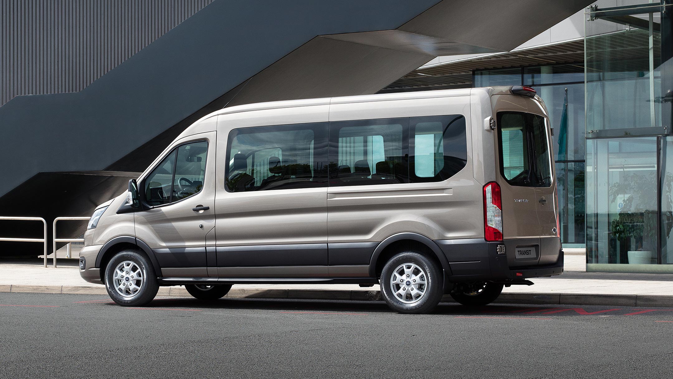 Ford Transit Kombi in Silber. Seitenansicht, parkend vor einem modernen Gebäude