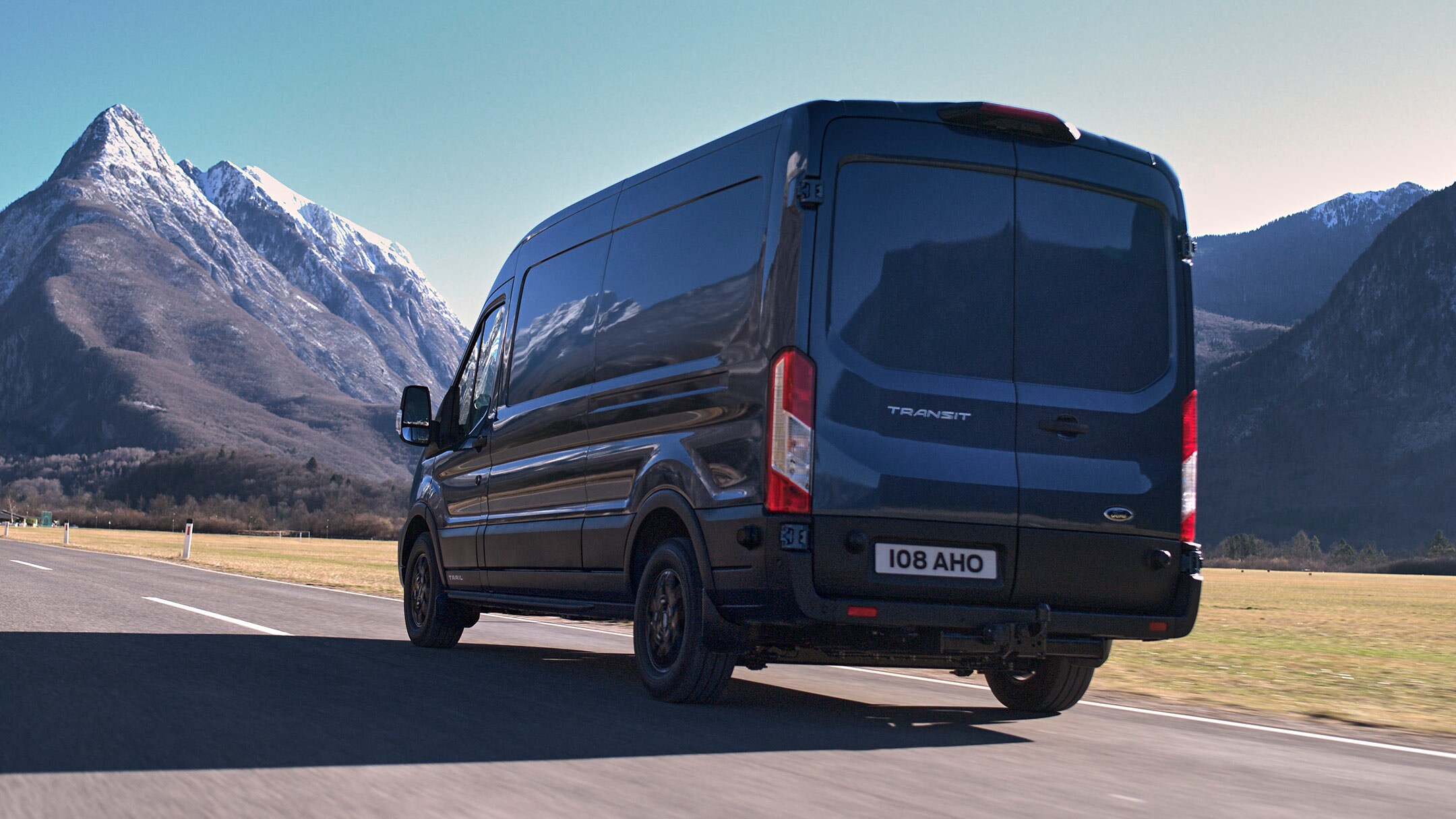 Ford Transit Kastenwagen in Blau. Dreiviertelansicht von hinten, fahrend auf einer Landstraße mit Bergen im Hintergrund