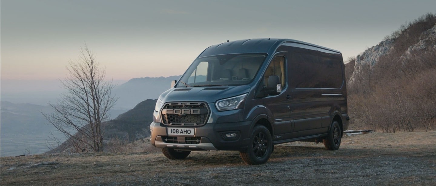 Ford Transit in Blau, fährt durch ländliches Gebiet mit Bergen im Hintergrund.