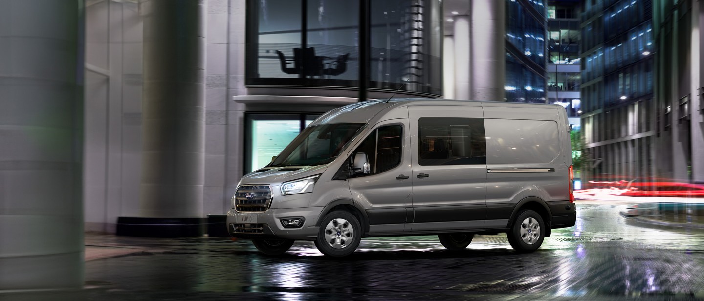 Ford Transit in Silber. Dreiviertelansicht, fahrend durch eine Stadt bei Nacht.