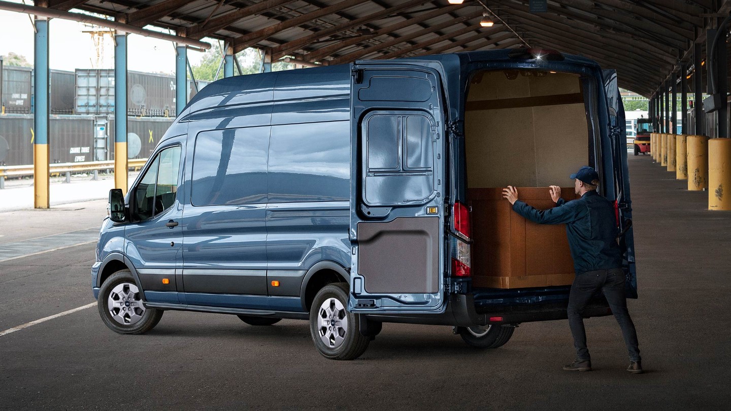 Ford Transit in Blau. Dreiviertelansicht von hinten, parkend unter einem Vordach mit weit geöffneten Hecktüren.