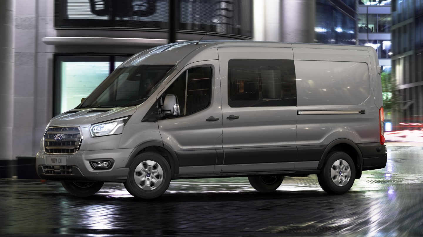 Ford Transit in Silber. Dreiviertelansicht, fahrend durch eine Stadt bei Nacht.